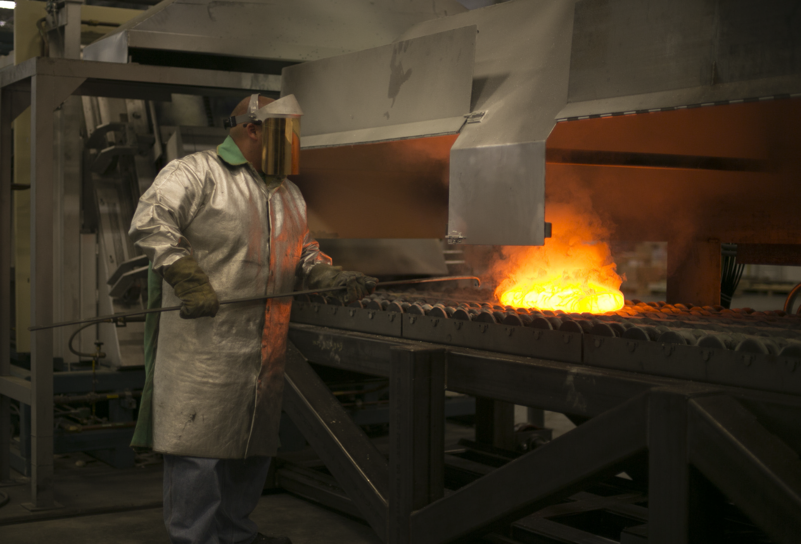 Industrial worker monitoring heated metal moving through a furnace on a conveyor system
