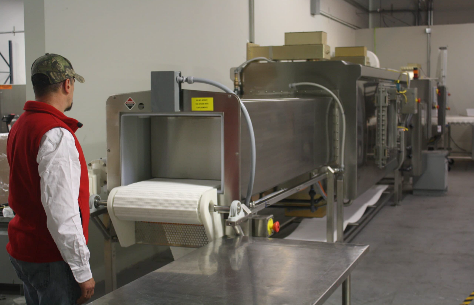 Operator standing beside a conveyor-fed industrial processing machine with metal housing